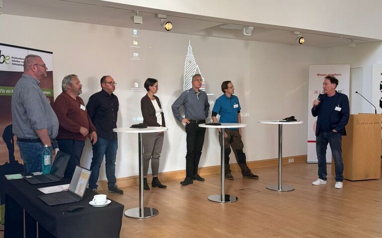 Foto der Veranstaltung. Sieben Personen stehen nebeneinander im Raum, eine Person spricht in ein Mikrofon. Vor ihnen stehen Stehtische, im Hintergrund sind Banner und eine Projektion an der Wand.