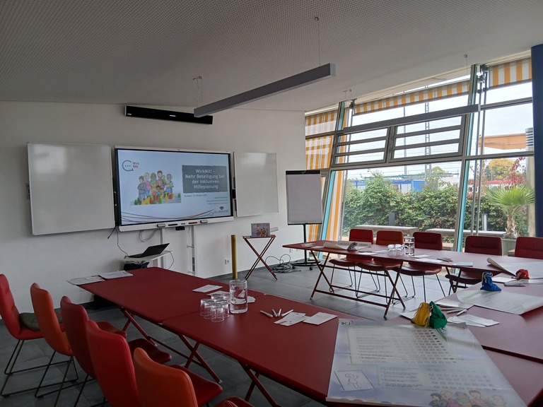 Seminarraum mit roten Stühlen und Konferenztischen, Präsentation zum Thema ‚WirkMit! – Mehr Beteiligung bei der inklusiven Hilfeplanung‘ auf großem Bildschirm, vorbereitete Arbeitsmaterialien und Flipchart, Blick nach außen durch große Fenster.