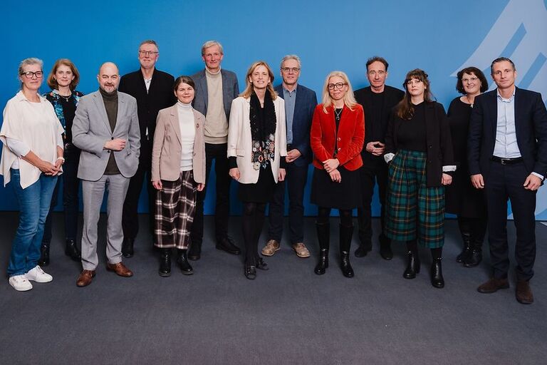 Eine Gruppe von dreizehn Personen steht in zwei Reihen vor einer blauen Wand mit einem stilisierten Bundesadler. Die Personen sind gemischt gekleidet – von leger bis formell – und blicken lächelnd in die Kamera. In der vorderen Reihe stehen sieben Personen, in der hinteren sechs. Die Gruppe posiert für ein offizielles Gruppenfoto in einem Innenraum mit grauem Teppichboden. © Jens Ahner/ BMBFSFJ