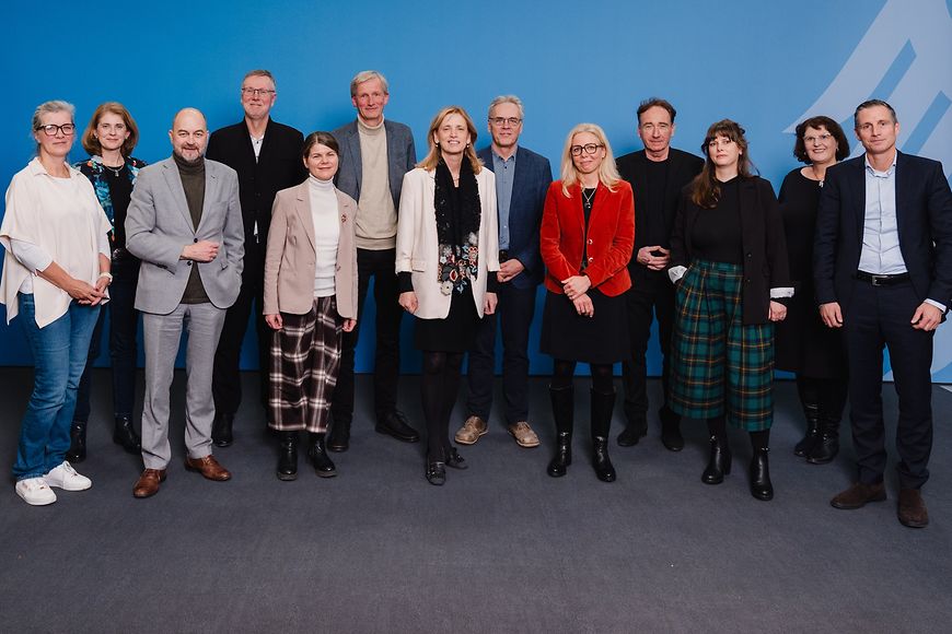 Eine Gruppe von dreizehn Personen steht in zwei Reihen vor einer blauen Wand mit einem stilisierten Bundesadler. Die Personen sind gemischt gekleidet – von leger bis formell – und blicken lächelnd in die Kamera. In der vorderen Reihe stehen sieben Personen, in der hinteren sechs. Die Gruppe posiert für ein offizielles Gruppenfoto in einem Innenraum mit grauem Teppichboden. © Jens Ahner/ BMBFSFJ